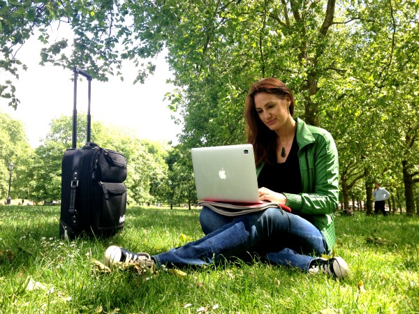 Natalie Sisson working on her laptop in a park, wheelie suitcase beside her